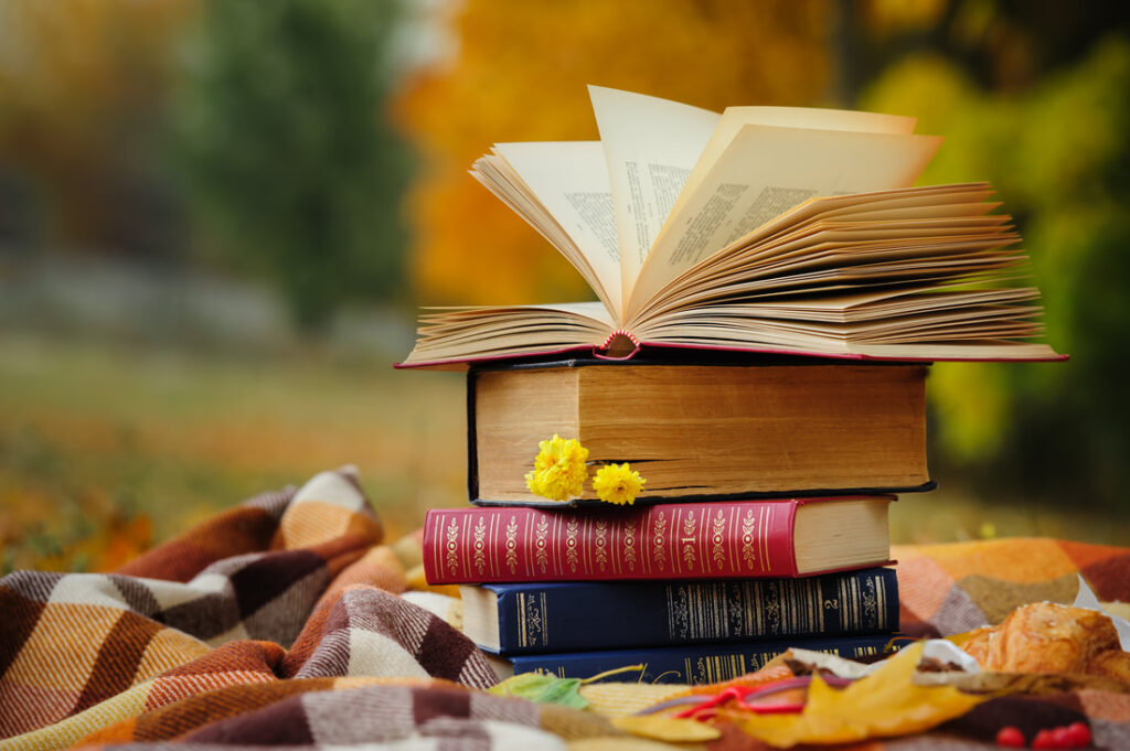 a stack of books on a picnic blanket. the top book is open and its pages flap in the breeze