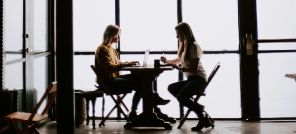 Two writers sit at a table by a large glass window