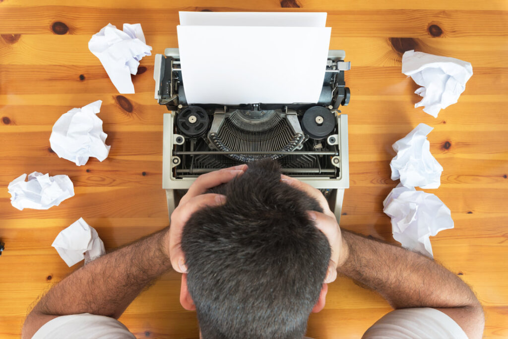 writer at a typewriter with head in hands surrounded by crumpled pages