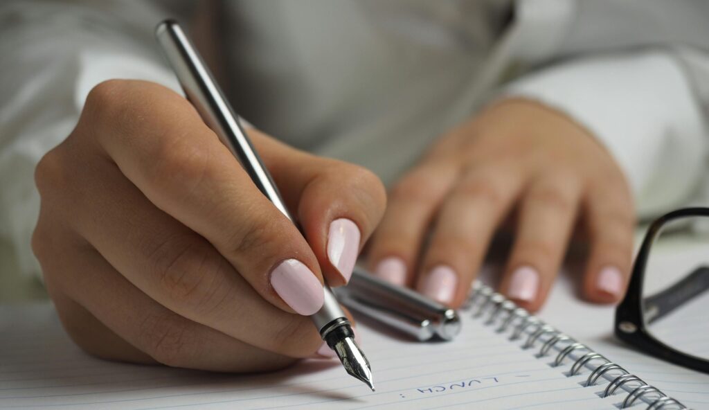 Woman in White Long Sleeved Shirt Holding a Pen Writing on a Paper