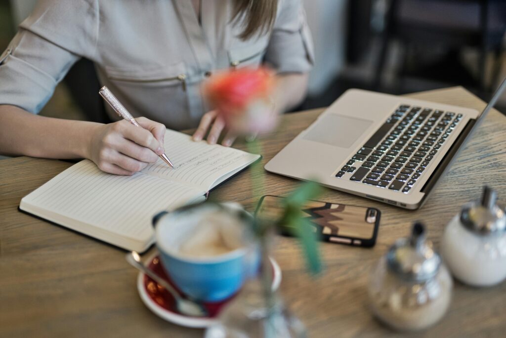 Author writing in notepad with laptop open on the desk and an empty cup of coffee