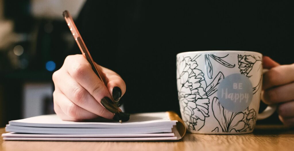 A writer at a table with a mug that reads BE HAPPY, handwriting in a notebook