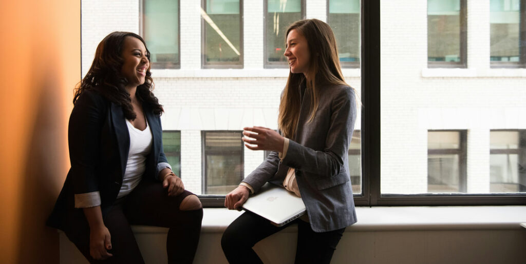 Two women sit in a stairwell window smiling and chatting. One has an open notepad on her lap.
