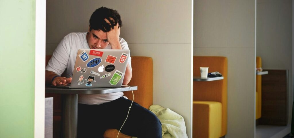 Writer sits alone with his laptop in a cafe booth. His head is in his hands and he looks stressed.